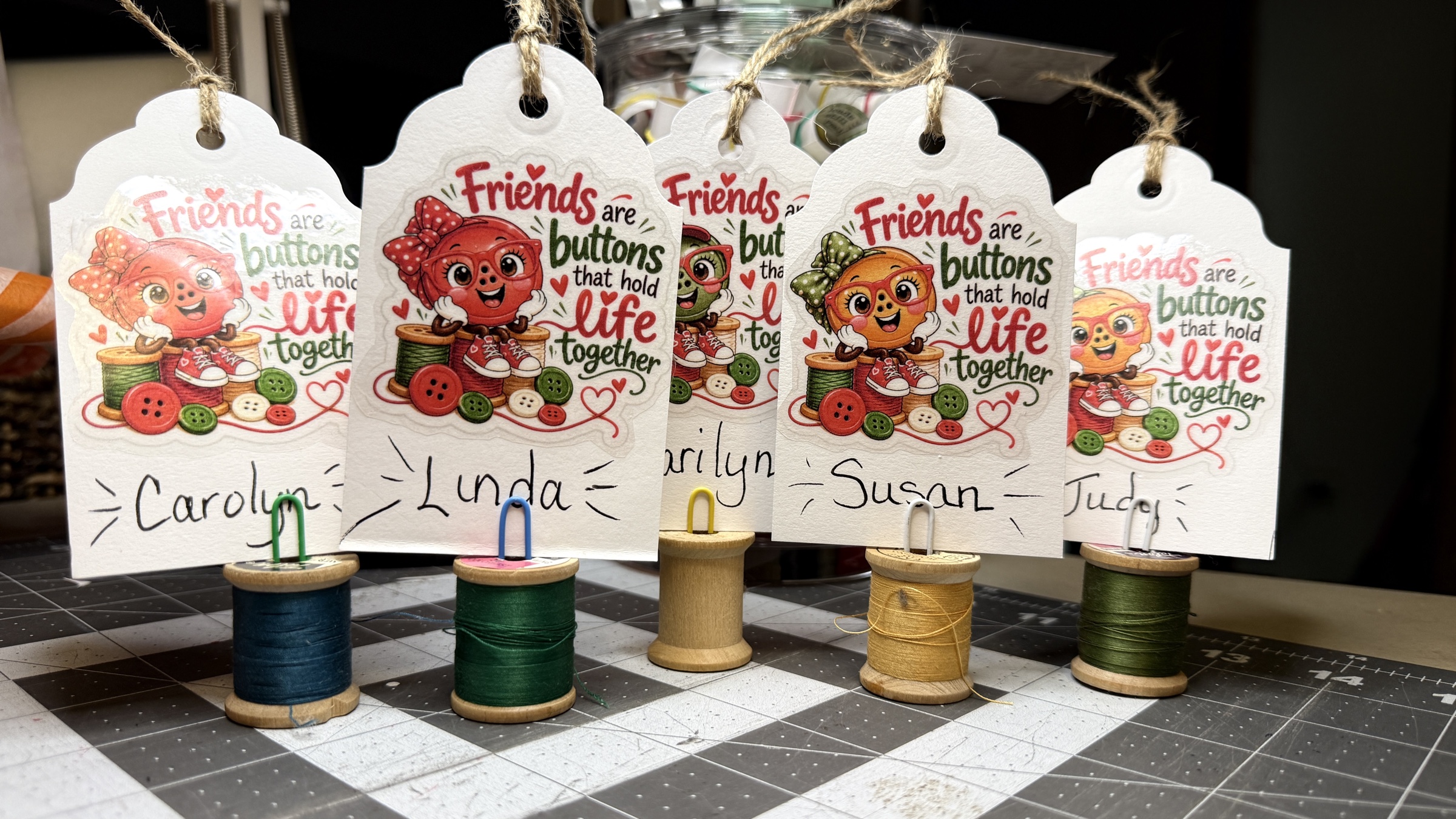 Five colorful friendship name tags hang from twine and are propped on thread spools with paper clips.