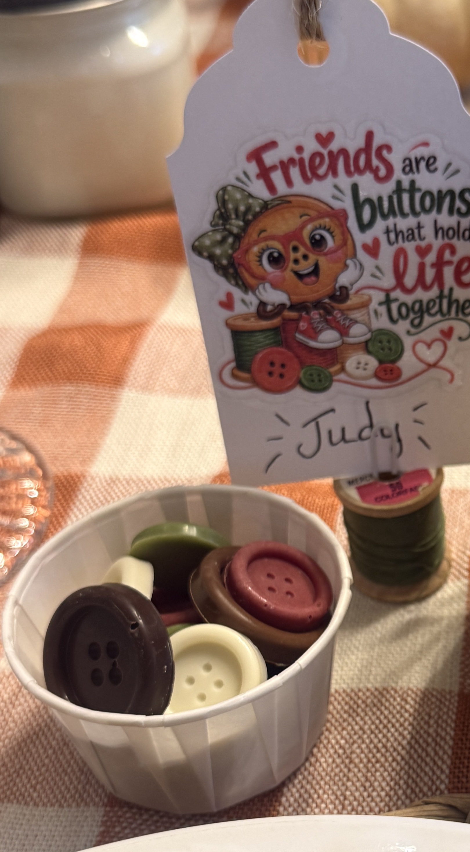A colorful friendship tag with an illustrated button character stands behind a cup of chocolate buttons on a checked tablecloth.